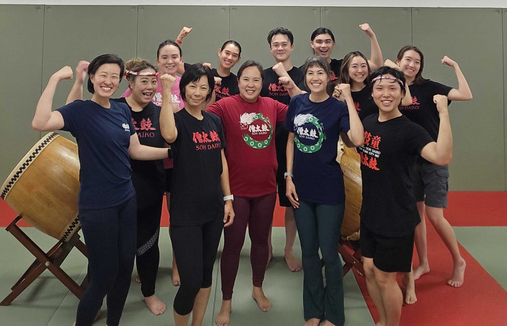 The current members of Soh Daiko pose as a group with their arms raised in victory.